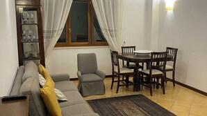 A living room with a sofa, armchairs, a dining table with chairs, a cabinet with glassware, and a window with curtains.