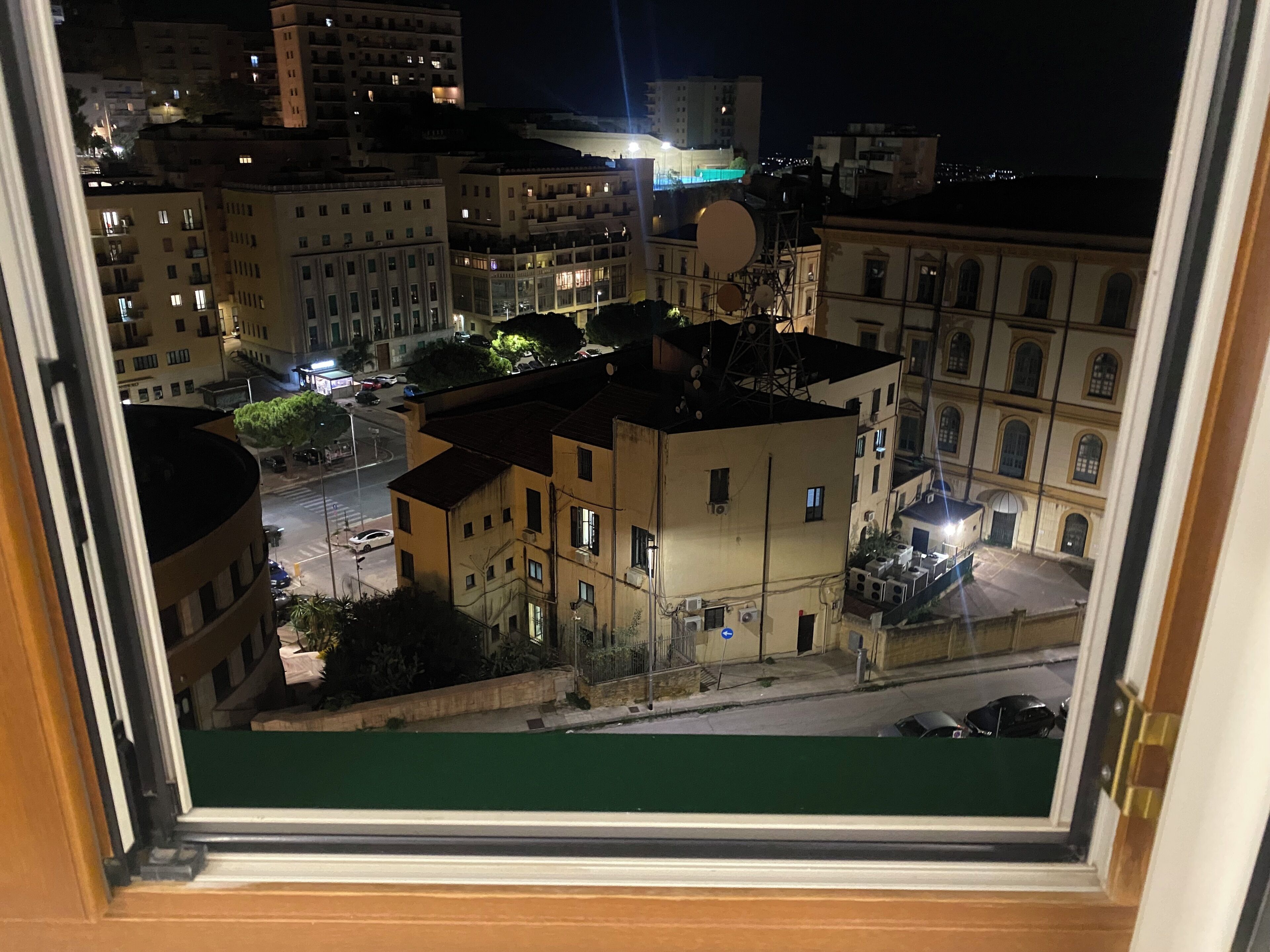 Une vue nocturne d’une ville, prise à travers une fenêtre, montrant des immeubles avec des balcons, une rue avec des voitures et une antenne parabolique bien visible.