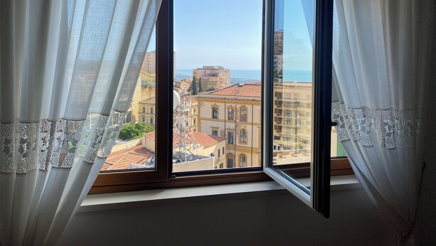 A room with a large window, white curtains, and a view of buildings and the sea.