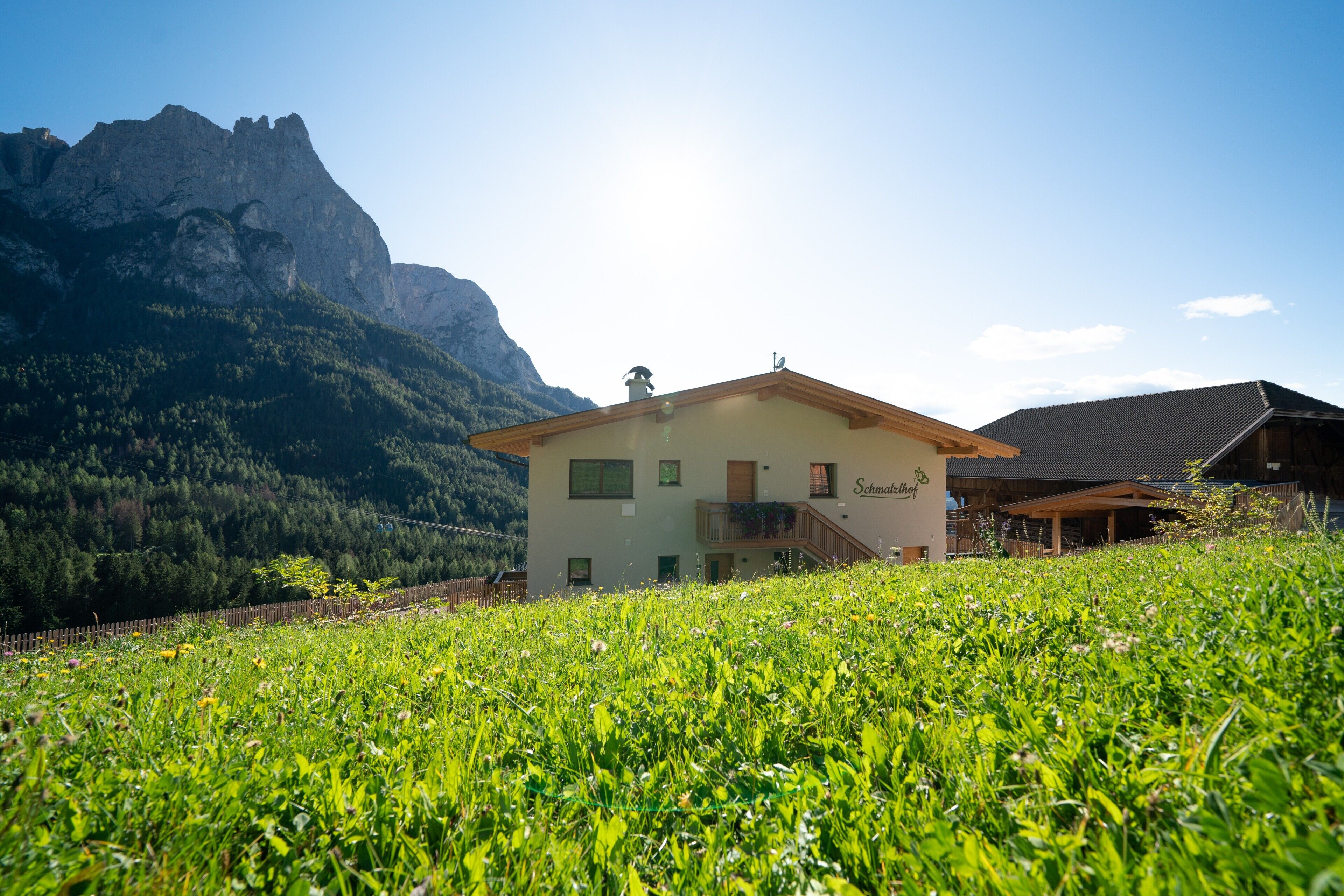 A mountain house with a balcony, surrounded by lush greenery and a mountain backdrop.