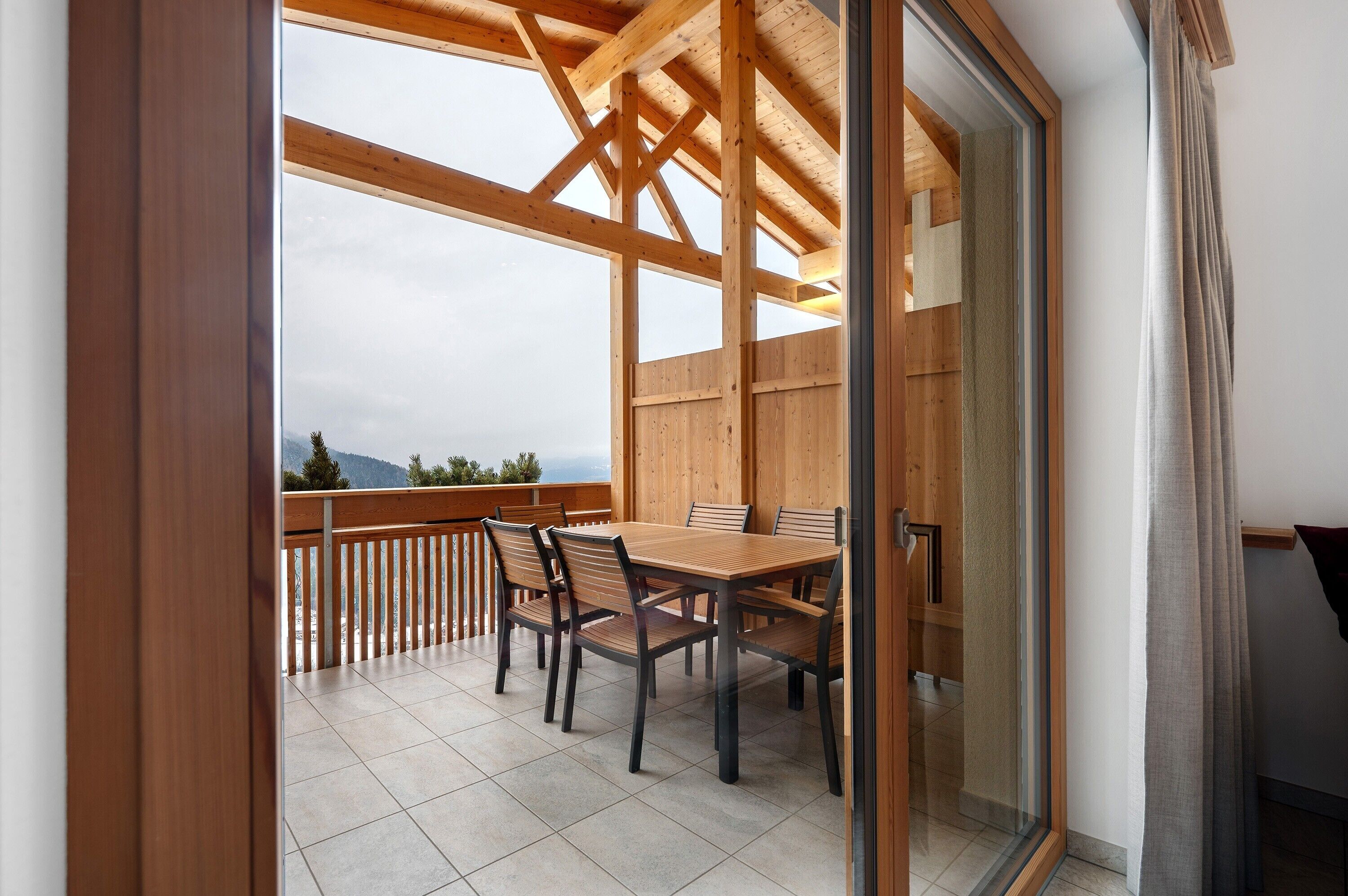 A wooden balcony with a table and chairs, a glass door, and a view of the outdoors.