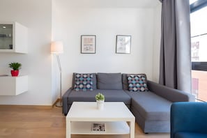 Una sala de estar moderna con un sofá gris, una mesita de centro blanca y una pequeña planta en maceta. Hay dos cuadros en la pared, una lámpara de pie y una ventana con cortinas.