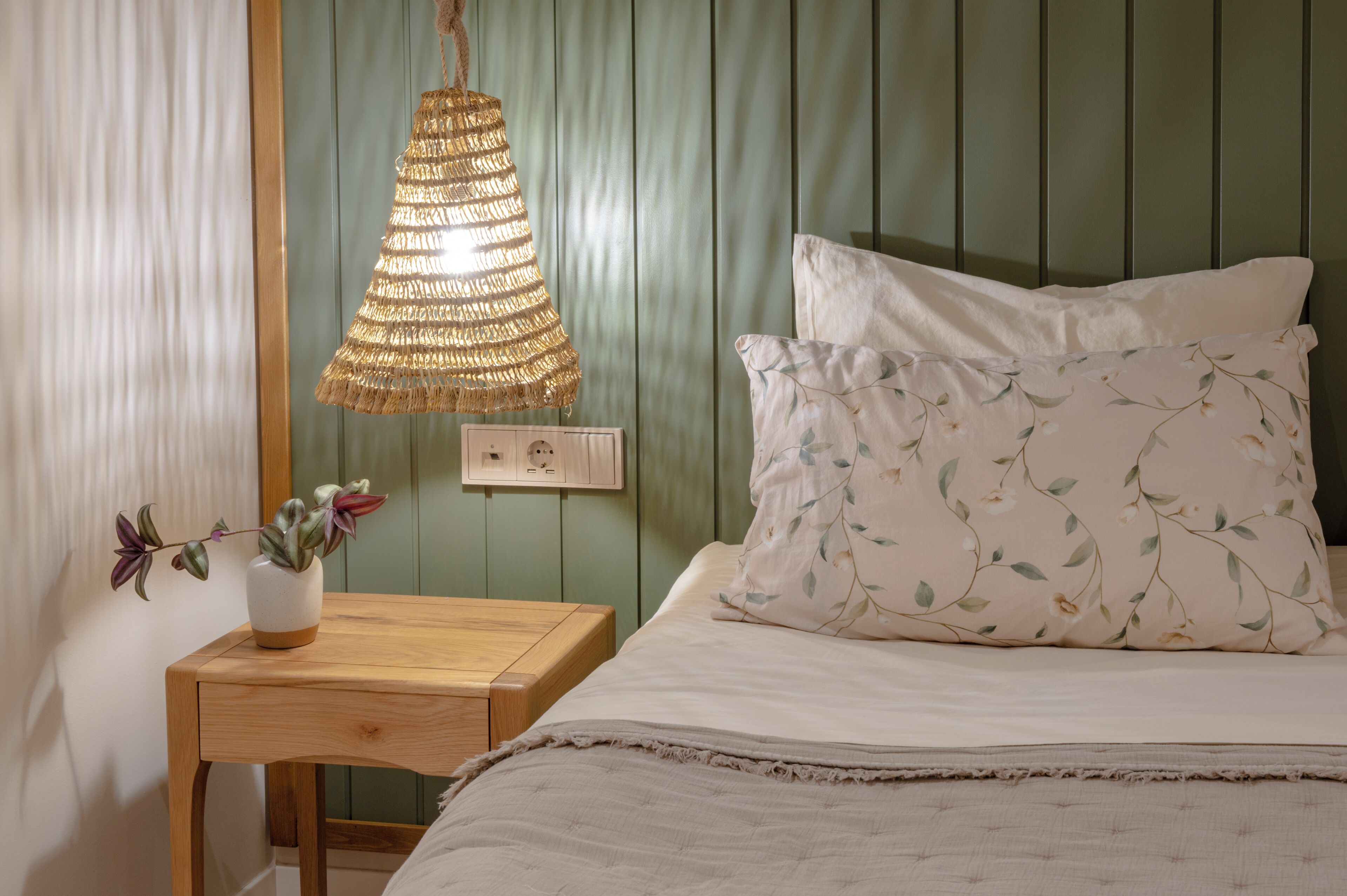 A bedroom with a bed, a bedside table, a hanging lamp, and a wall socket.