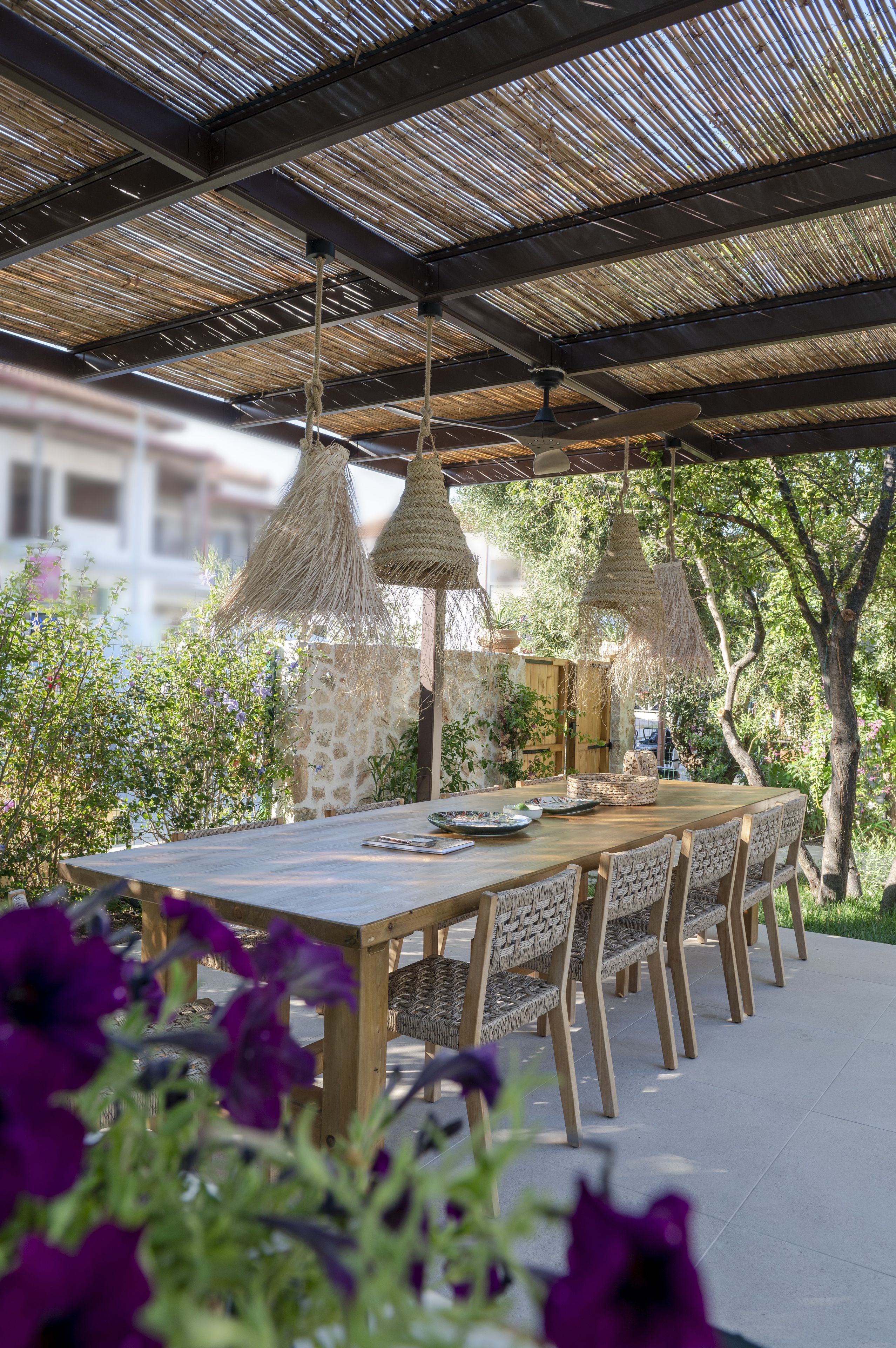 An outdoor dining area with a long table and chairs, surrounded by greenery and a thatched roof structure.