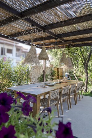 An outdoor dining area with a long table and chairs, surrounded by greenery and a thatched roof structure. - Elea 1 - Modern, comfortable furnishings, close to the beach (Ierissos)