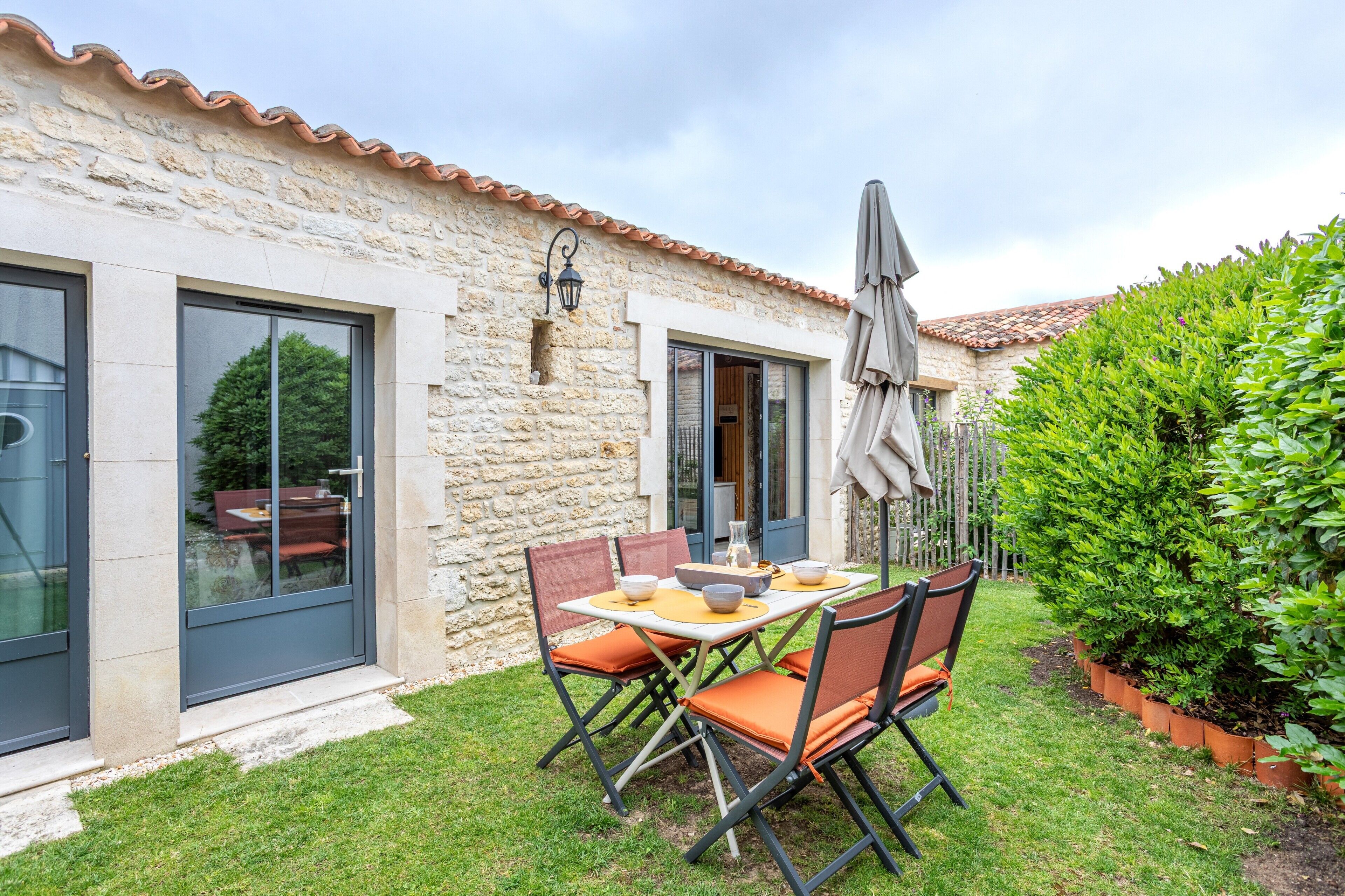 A stone building with a tiled roof, a patio with a table and chairs, and a well-maintained garden.