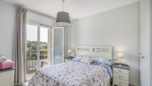 Un dormitorio con cama, dos mesitas de noche, una lámpara colgante, una ventana con cortinas y vistas al exterior.