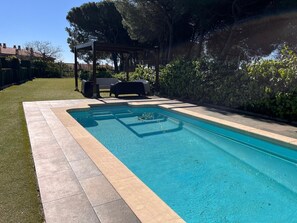 Una piscina con borde de azulejos, rodeada de césped y una pérgola con asientos.