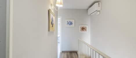 A hallway with a wooden floor, white walls, and a white railing. There are three framed pictures on the wall.