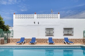 A pool with four blue lounge chairs and a white building with a decorative wall and windows.