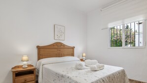 A bedroom with a wooden headboard, a bed with a floral bedspread, two bedside lamps, a small wooden nightstand with drawers, a framed picture on the wall, and a window with a view of greenery. - Holiday Home 'Manuel' with Shared Pool, Wi-Fi and Air Conditioning (Conil)