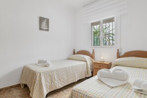 A bedroom with two beds, a nightstand, a window with a view of greenery, and a wall-mounted artwork.
