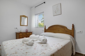A bedroom with a wooden bed, a bedside table with a lamp, a window with bars, and a framed picture on the wall.
