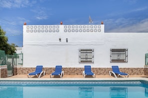 A pool with four blue lounge chairs and a white building with a decorative wall and windows.