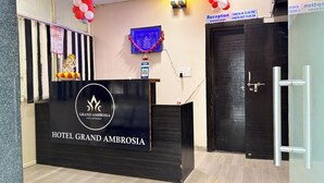 Ein Empfangsschalter mit einem Schild für das Hotel Grand Ambrosia, ein blauer dekorativer Gegenstand an der Wand und eine Tür mit Hinweisschild für Check-in/Check-out.