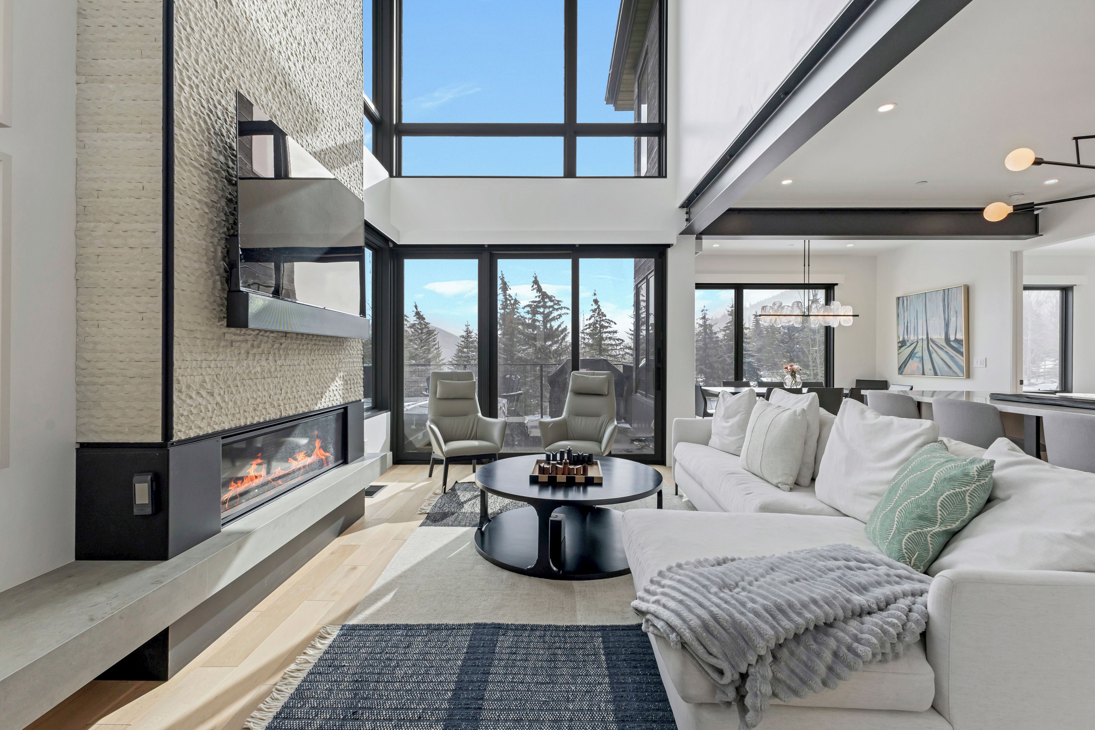 Un salon moderne avec une cheminée, un canapé, une table basse et une vue sur les arbres à travers de grandes fenêtres.