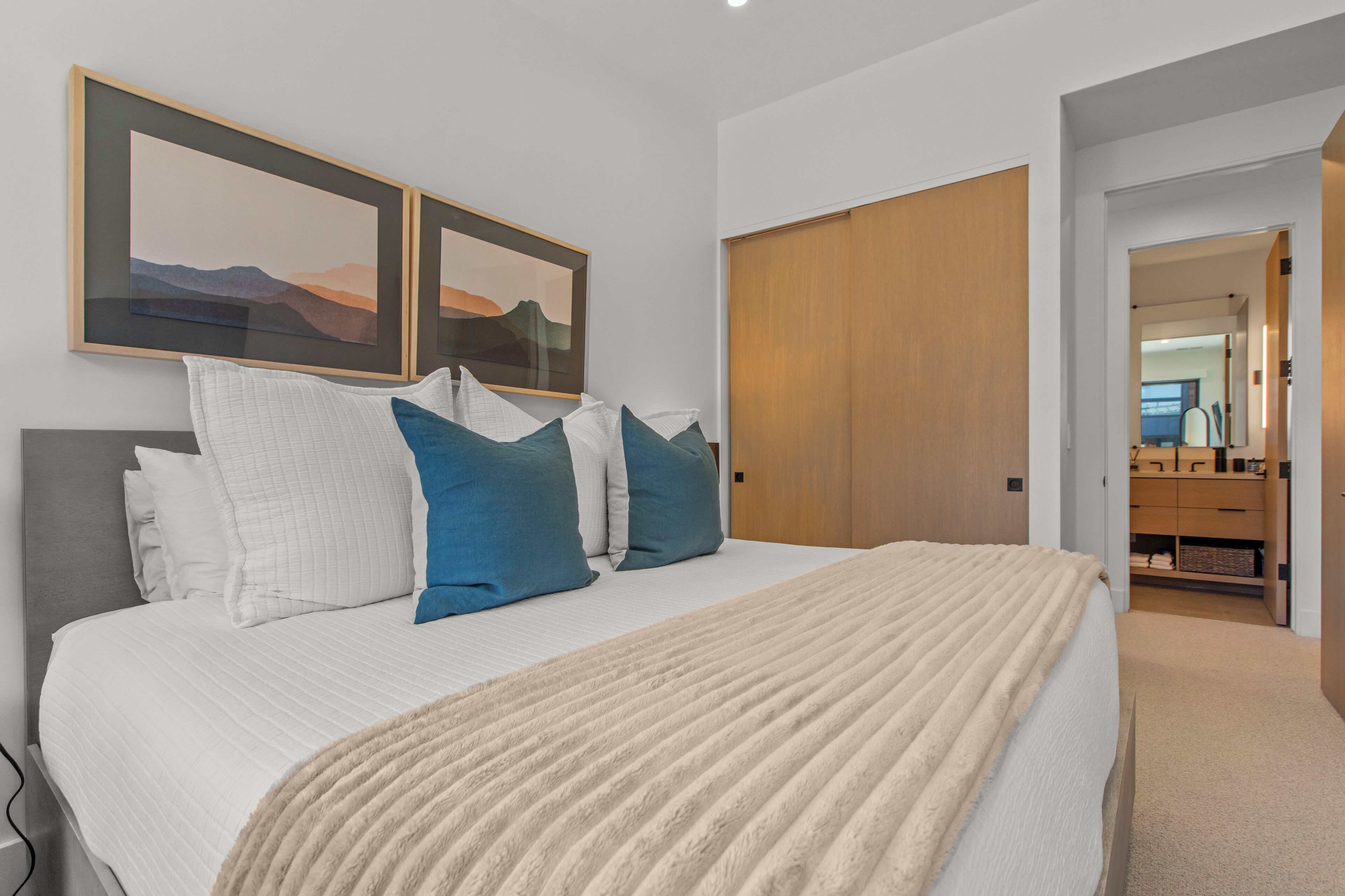 A bedroom with a large bed, a wooden wardrobe, and two framed landscape paintings on the wall.