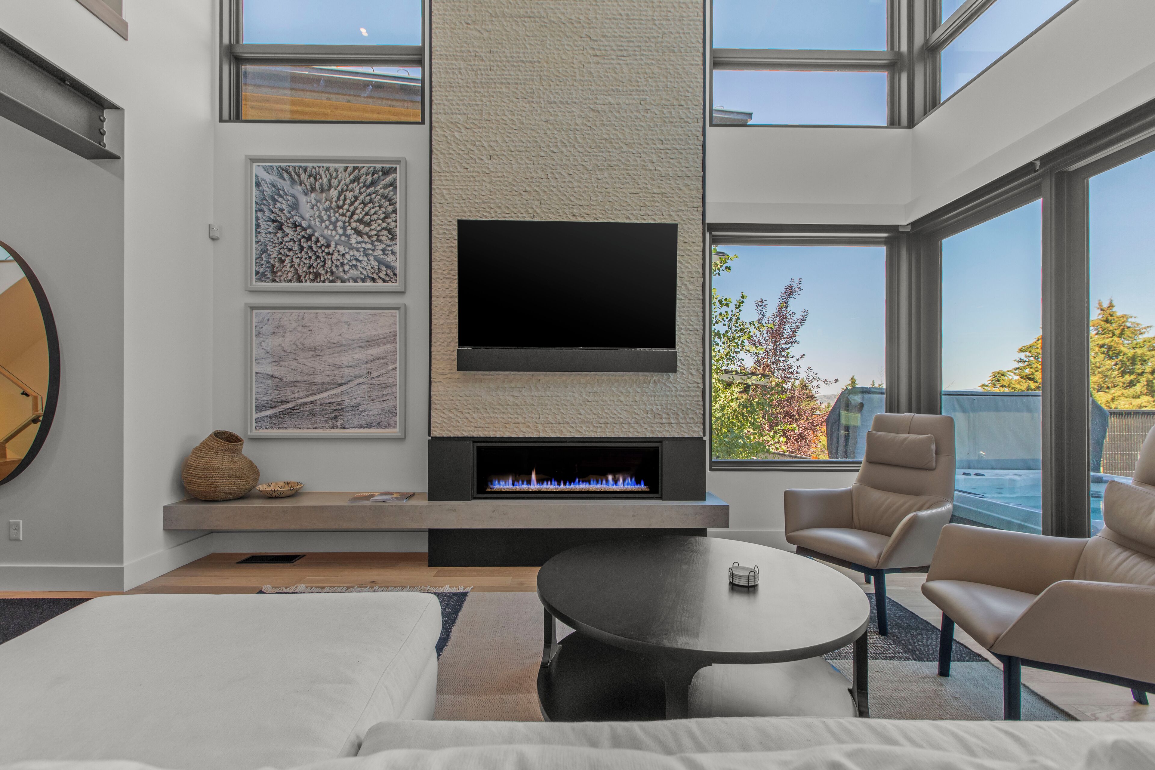 A modern living room with a fireplace, a flat-screen TV mounted on a textured wall, and a large window offering a view of the outdoors.