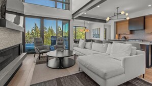 A spacious living area with a large sofa, two armchairs, and a coffee table. There is a fireplace, large windows offering a view of the outdoors, and a kitchen area visible in the background. - Modern Home in Canyons Village by InvitedHome | Hot Tub, Game Room, Hike, Bike (Park City)