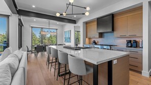 A modern kitchen with a central island, bar stools, and a dining area with a view. - Modern Home in Canyons Village by InvitedHome | Hot Tub, Game Room, Hike, Bike (Park City)