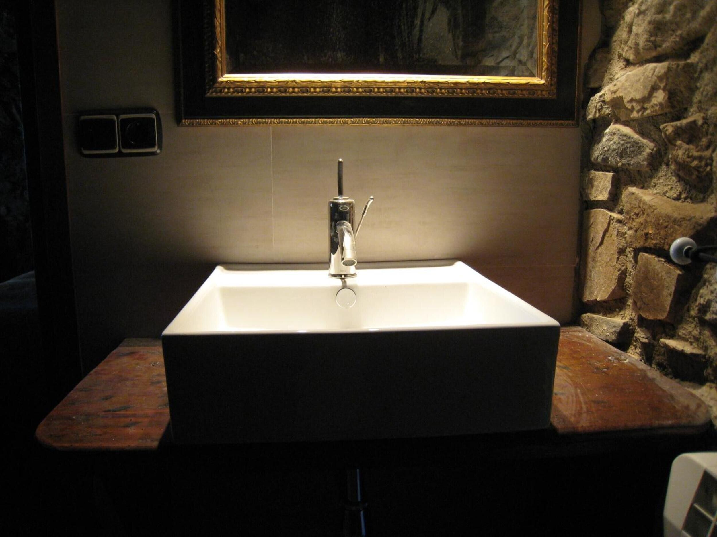Un baño con lavamanos blanco, pared de piedra y una obra enmarcada sobre el lavamanos.
