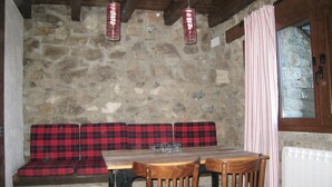 Un mur de pierres, une table en bois avec deux chaises, un banc à carreaux rouges et une fenêtre avec des rideaux.