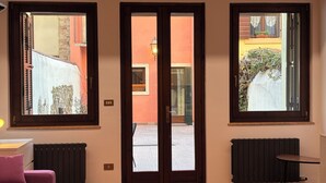 Quarto com sofá rosa, radiador e uma mesa pequena. Três janelas oferecem vista para o exterior.