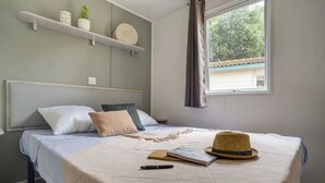 Ein ordentlich bezogenes Bett mit Hut, Buch und Stift auf der Tagesdecke. Kissen liegen auf dem Bett, ein Regal mit Dekoration und ein Fenster mit Vorhängen.