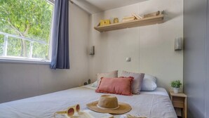 A neatly made bed with pillows, a hat, sunglasses, and a book on a white bedspread. There is a window with a blue curtain, a shelf with books and a plant, and a wall-mounted light.