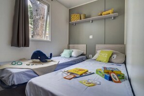Ein Hotelzimmer mit zwei Betten, einem Fenster mit Vorhängen, einem Regal mit Dekoration und Spielzeug sowie Büchern auf den Betten.