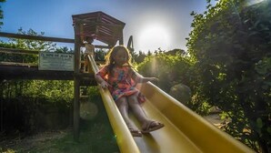 Un niño resbalando por un tobogán amarillo en un parque infantil con una estructura de madera y un cartel al fondo.