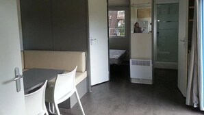 Une petite pièce comprenant un lit, une petite table et une chaise. Une porte donne accès à une autre pièce qui contient une fenêtre et un réfrigérateur.