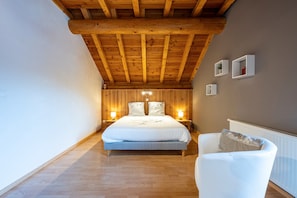 Een slaapkamer met een houten plafond, een groot bed, nachtkastjes, een witte stoel en een radiator.