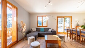 En moderne stue med en hjørnesofa, et spisebord med stole og et stort vindue med udsigt over et sneklædt landskab.