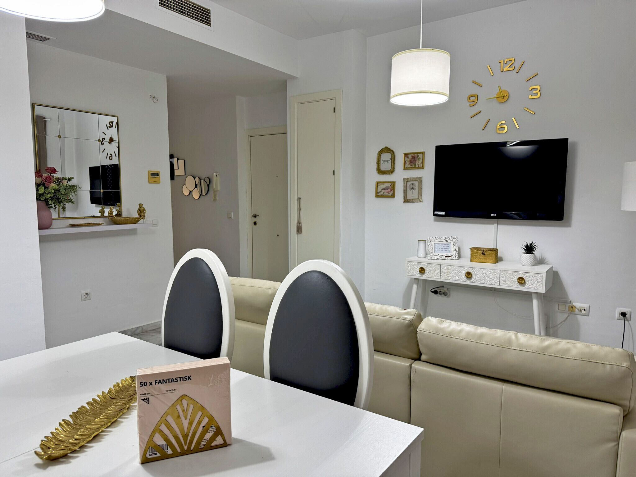 Una sala de estar moderna con un sofá blanco, una mesa de comedor con dos sillas, un televisor montado en la pared y un reloj decorativo.