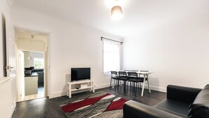 A modern living room with a dark wood floor, a black leather sofa, a TV on a white stand, a dining area with a table and chairs, and a kitchen visible through an open doorway.