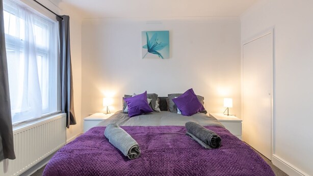A bedroom with a large bed, purple bedding, two nightstands with lamps, a window with curtains, and a wall-mounted artwork.