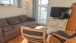 A living room with a sofa, two wooden chairs, a small round table with a candle, a television, and a view of a body of water through a large window. - Hausboot in Kroeslin am Bodden (Kröslin)