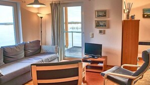 A modern living room with a sofa, a wooden chair, a TV, and a view of a body of water through the window. - Hausboot in Kroeslin am Bodden (Kröslin)