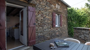 Ein Steinhaus mit einer durchgehenden Tür erkennbarer Küche, einem Tisch mit Buch und Sonnenbrille sowie einer Steinwand mit Grünpflanzen.