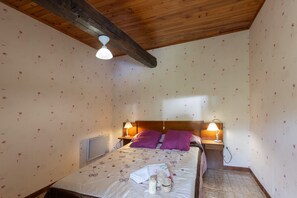 Ein Schlafzimmer mit Holzboden, blumigem Wandpapier, einem Bett mit violetten Kissen, zwei Nachttischlampen und einem kleinen Nachttisch.
