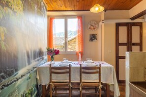 Un coin repas avec une table dressée pour deux personnes, des chaises en bois, une horloge au mur et une vue sur un paysage montagneux par la fenêtre.