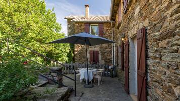 Un bâtiment en pierre avec une table et des chaises sur une terrasse, une clôture en bois et un grand parasol.