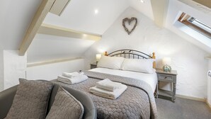 Un dormitorio con techo inclinado, una cama con ropa de cama blanca, una decoración con forma de corazón en la pared y una mesita de noche con una lámpara.