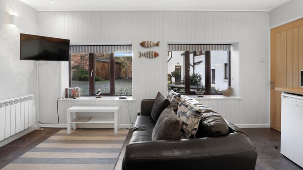 Una sala de estar moderna con una pared blanca con paneles, un televisor de pantalla plana montado sobre un banco blanco, un sofá de cuero con cojines estampados y vistas al exterior a través de amplias ventanas.