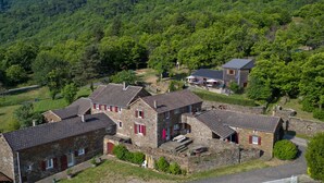A stone building with a tiled roof, surrounded by greenery and a forested area.