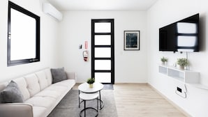 Ein modernes Wohnzimmer mit einer Couch, einem runden Tisch mit Pflanze, einem an der Wand montierten Fernseher und einem großen Fenster.