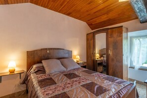 A bedroom with a wooden ceiling, a bed with a patterned quilt, bedside lamps, a wardrobe, and a window with curtains. - Holiday Home 'La Grange' with Mountain View, Shared Pool and Air Conditioning (Gravières)