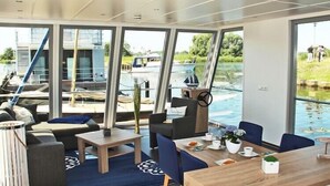 A modern interior with large windows, a dining area, and a view of the water outside. - Hausboot in Ueckermuende (Ueckermünde)