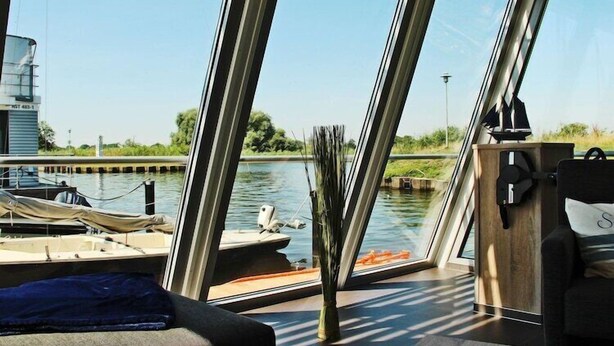 A modern boat interior with large windows, a wooden deck, and a view of a canal with boats. - Hausboot in Ueckermuende (Ueckermünde)